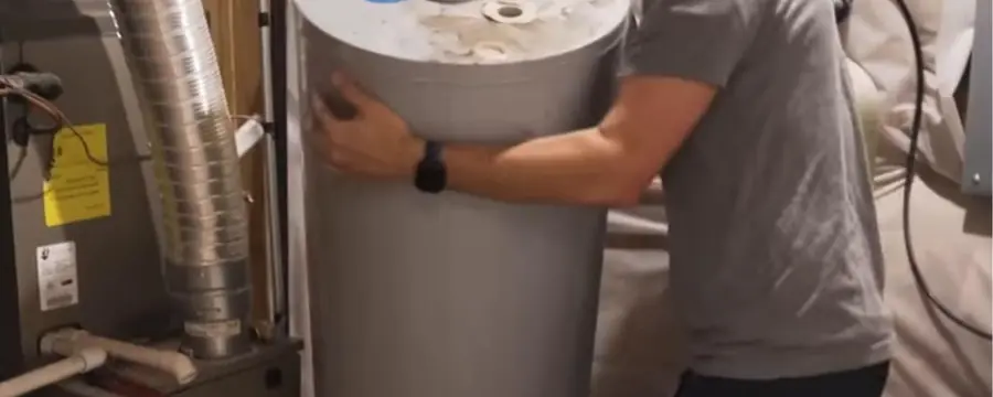A technician carries a new tank water heater into a utility space beside a furnace and ductwork.